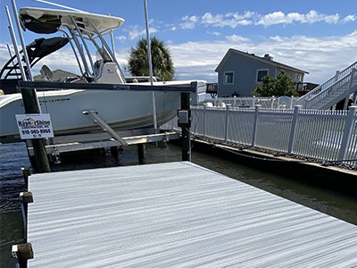 gray-blue dock with railing and a medium sized boat with water, sky, and a house in background