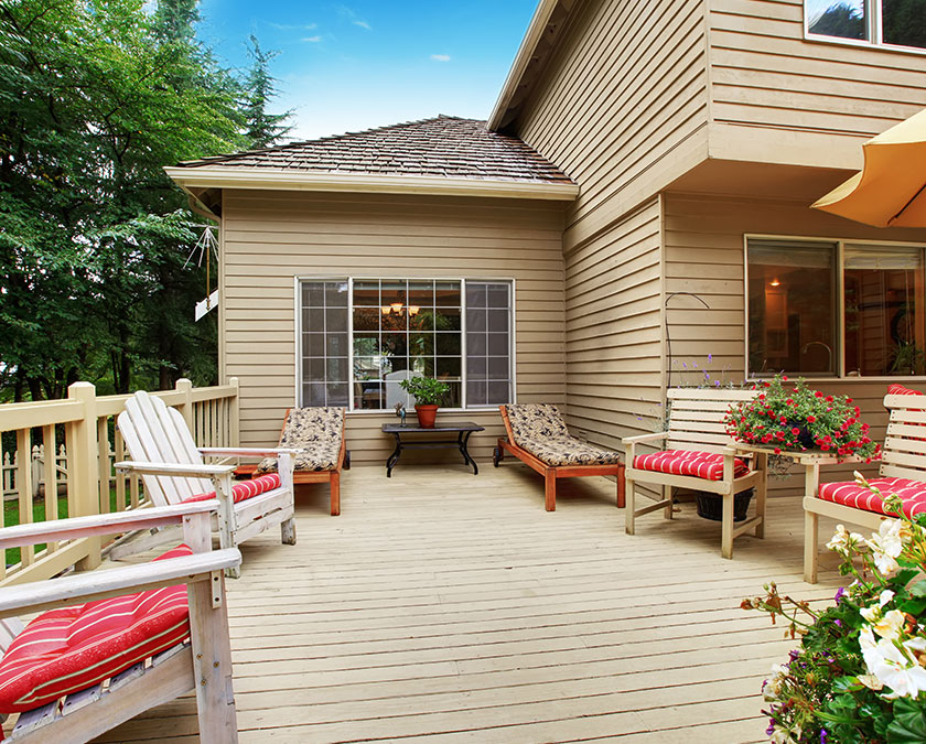 light brown house and deck and railing with outdoor wooden furniture set, red-with-white-stripes pillows on furniture, some outdoor pot plants and trees in background