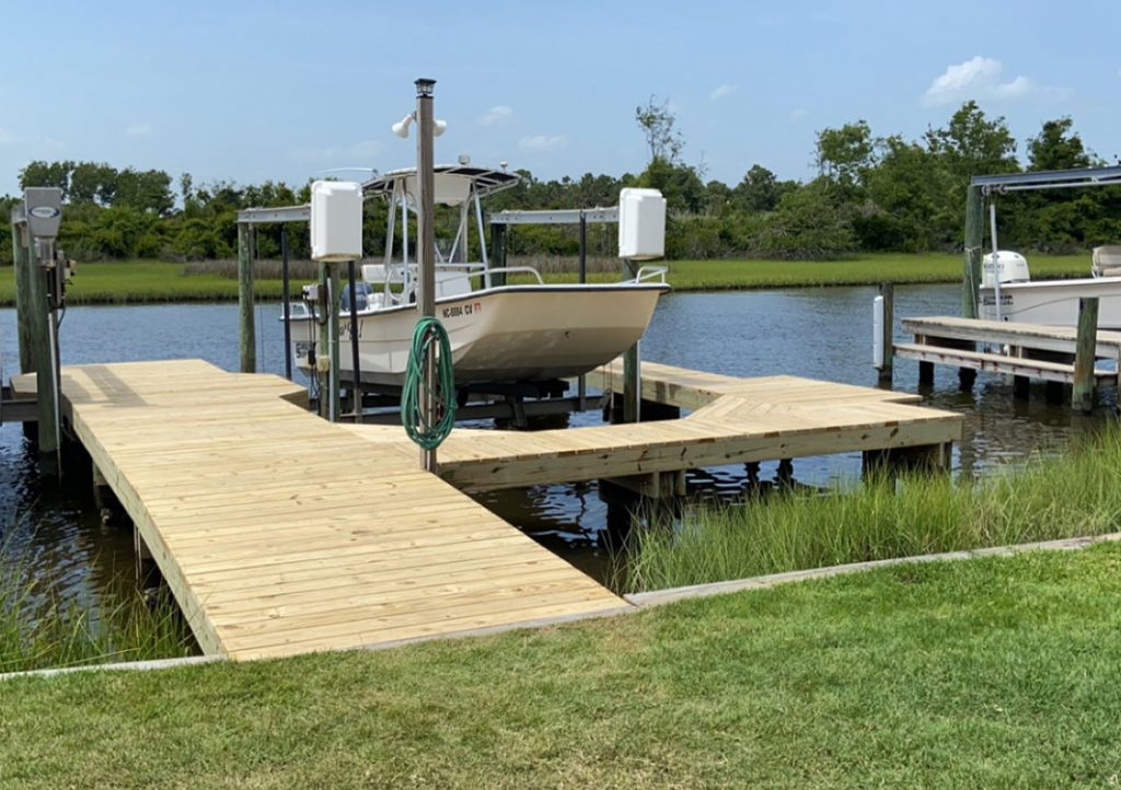 wooden dock with boat lift holding up medium sized boat, dock encompses boat partially, background is vegetation and water