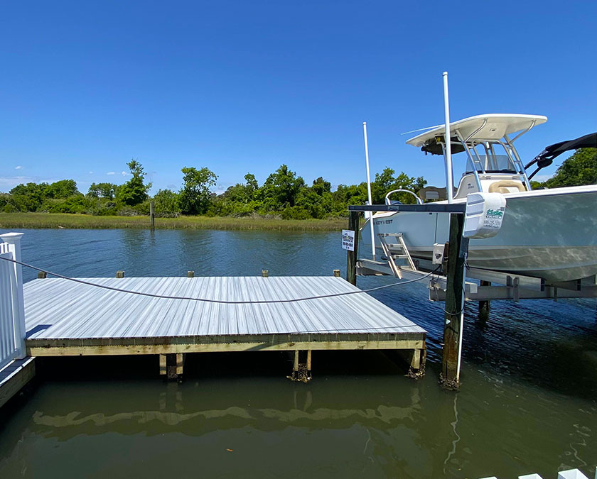 gray-blue dock with railings and a boat lift, lifting a medium sized boat out of water, water, field and dense vegetation in background