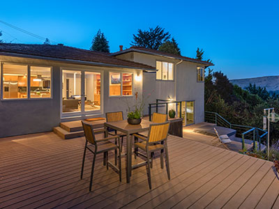 large composite deck with steps leading into backdoor, outdoor furniture set, metal railings along steps leading up to the deck, shrubs and trees in background with a house well-lit house in view