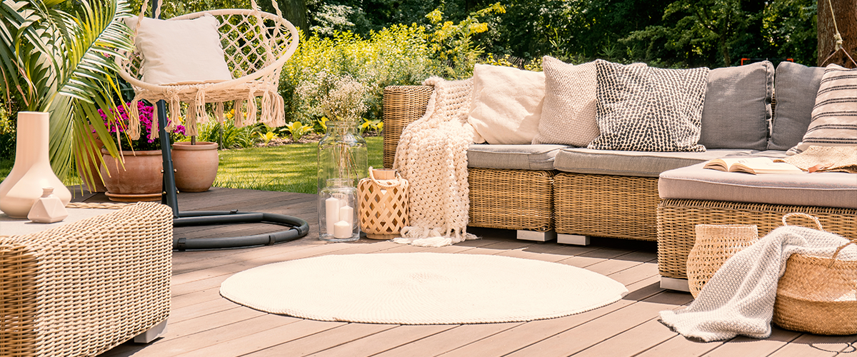 A cozy outdoor patio with a woven rattan sectional sofa, a hanging macrame chair, soft pillows and blankets, and lush green surroundings.