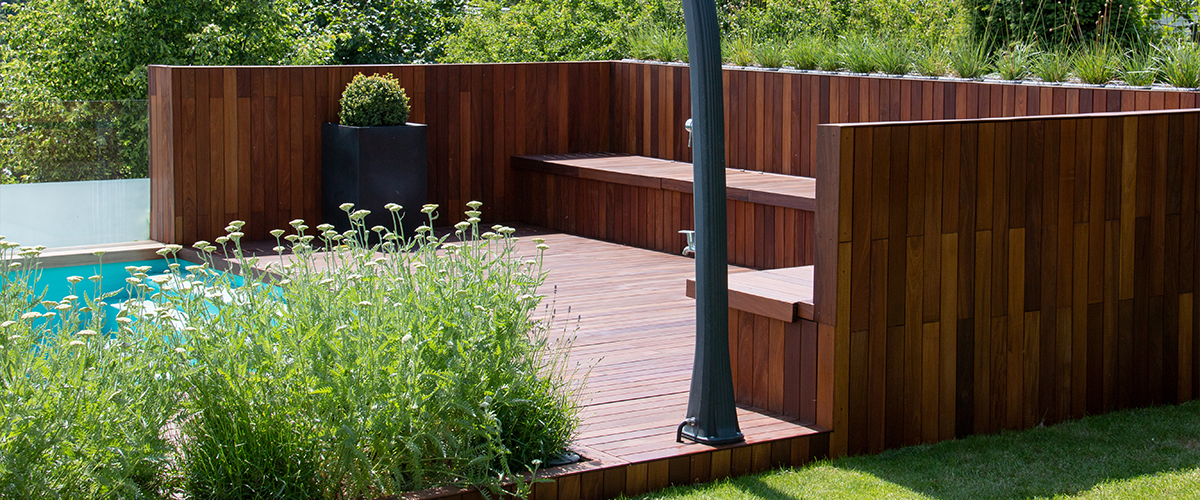 A modern backyard pool area featuring a sleek outdoor shower, dark wood decking, built in seating, and lush greenery.