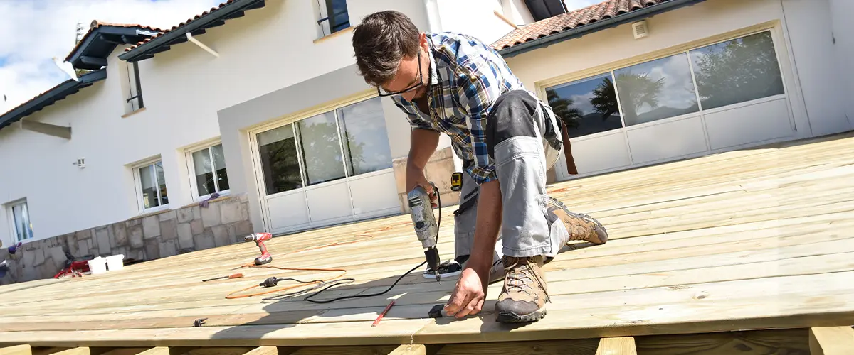 Carpenter building a wooden deck on a modern home exterior in Whispering Pines, using a power drill. Deck construction, home improvement.