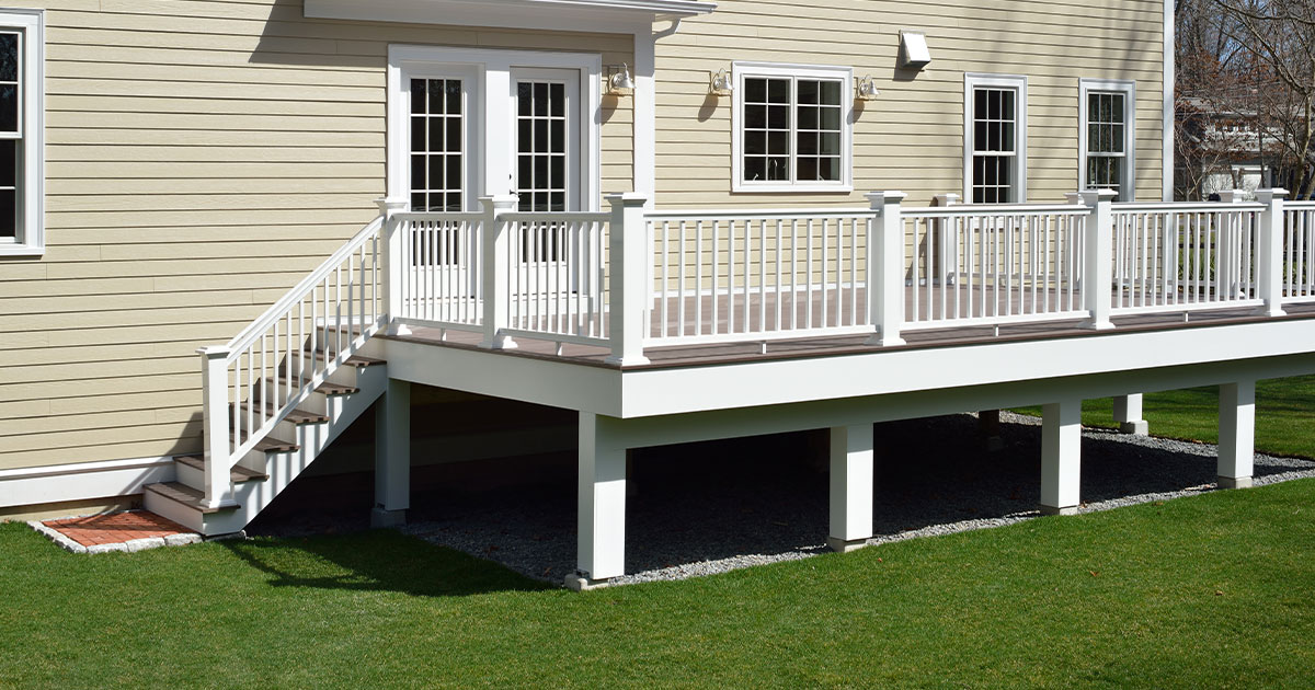 Elevated deck with gravel underneath, white railing attached to a beige house with French doors, featuring a staircase leading to the backyard lawn - modern home exterior