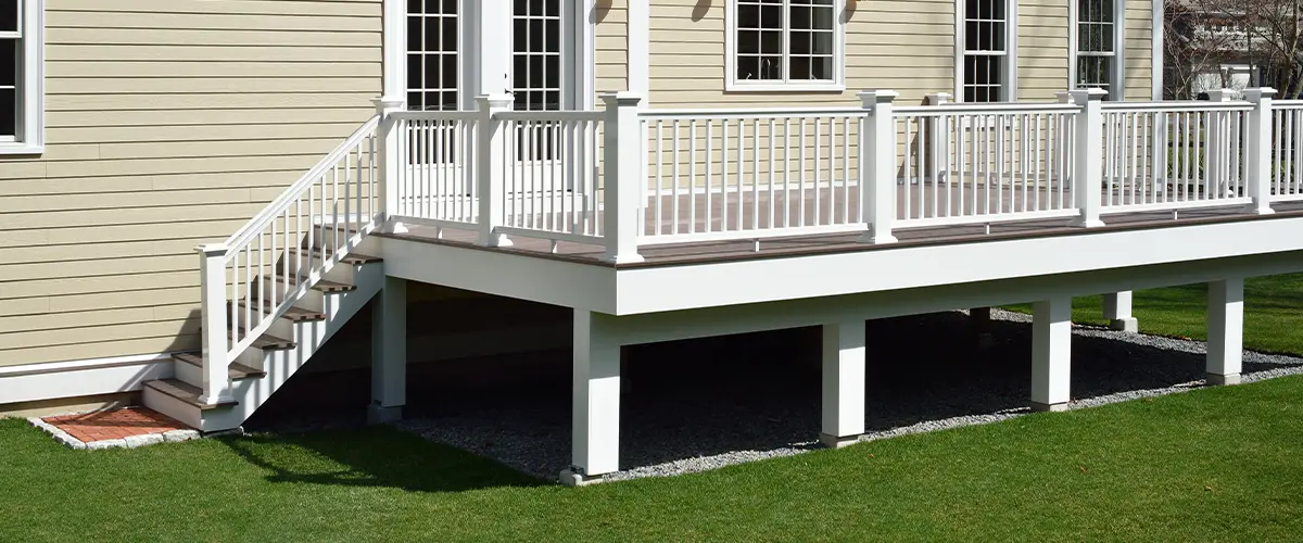 Elevated deck with gravel underneath, white railing attached to a beige house with French doors, featuring a staircase leading to the backyard lawn - modern home exterior