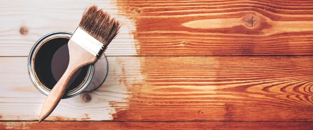 Paintbrush resting on an open can of wood stain with partially stained wooden planks