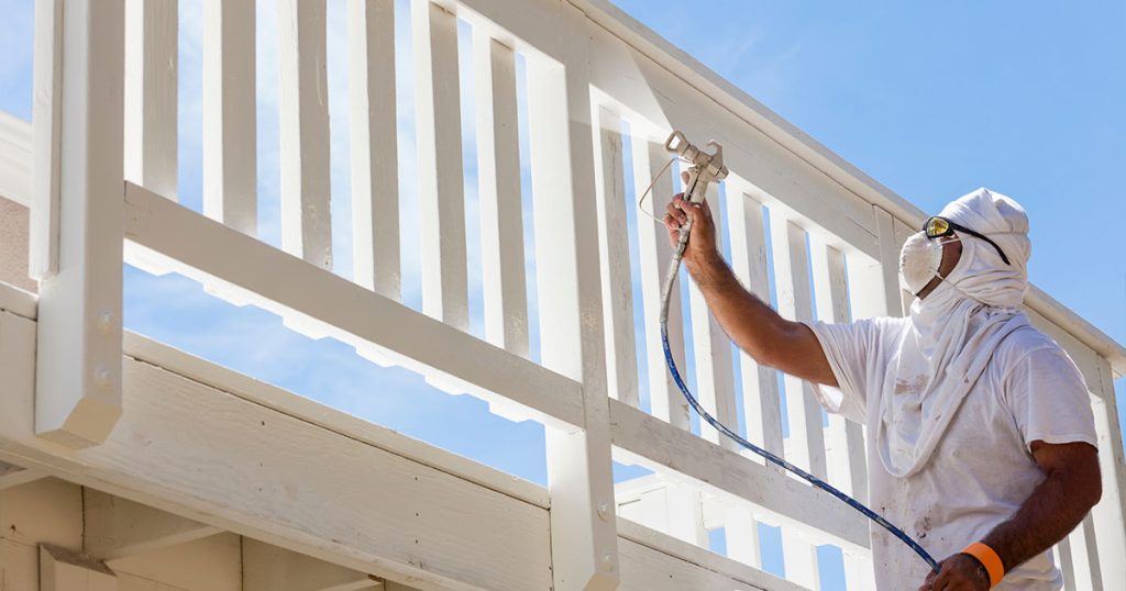Contractor spray painting white wood deck railing for refinishing and maintenance under bright blue sky.