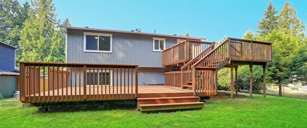 Spacious wooden backyard deck with stairs and railings, attached to a modern home, surrounded by green lawn and trees.
