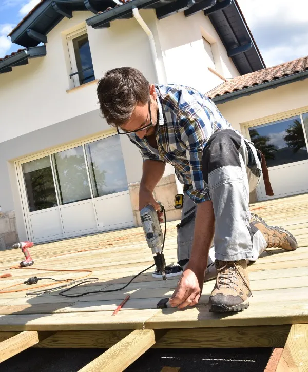 A man uses a drill to work on a wooden deck, focused on his task in a sunny outdoor setting