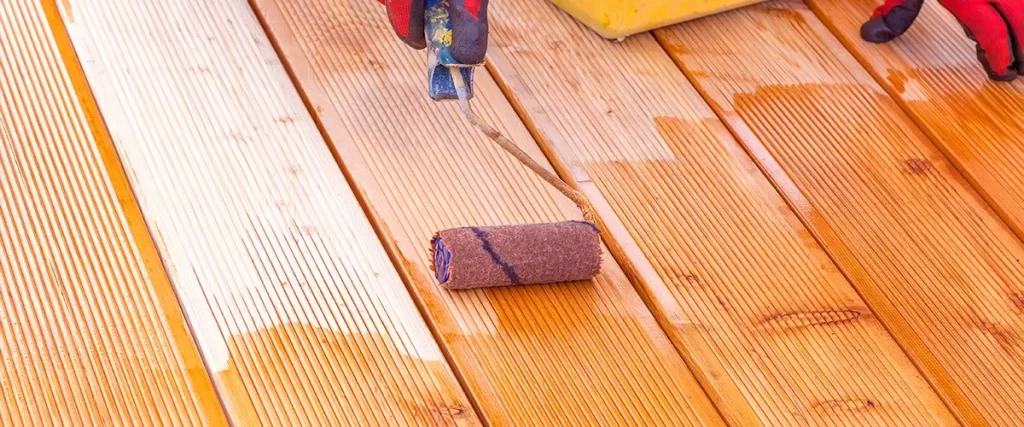 A close-up view of a person wearing red gloves using a paint roller to apply a fresh coat of orange-toned stain onto grooved wooden deck boards.