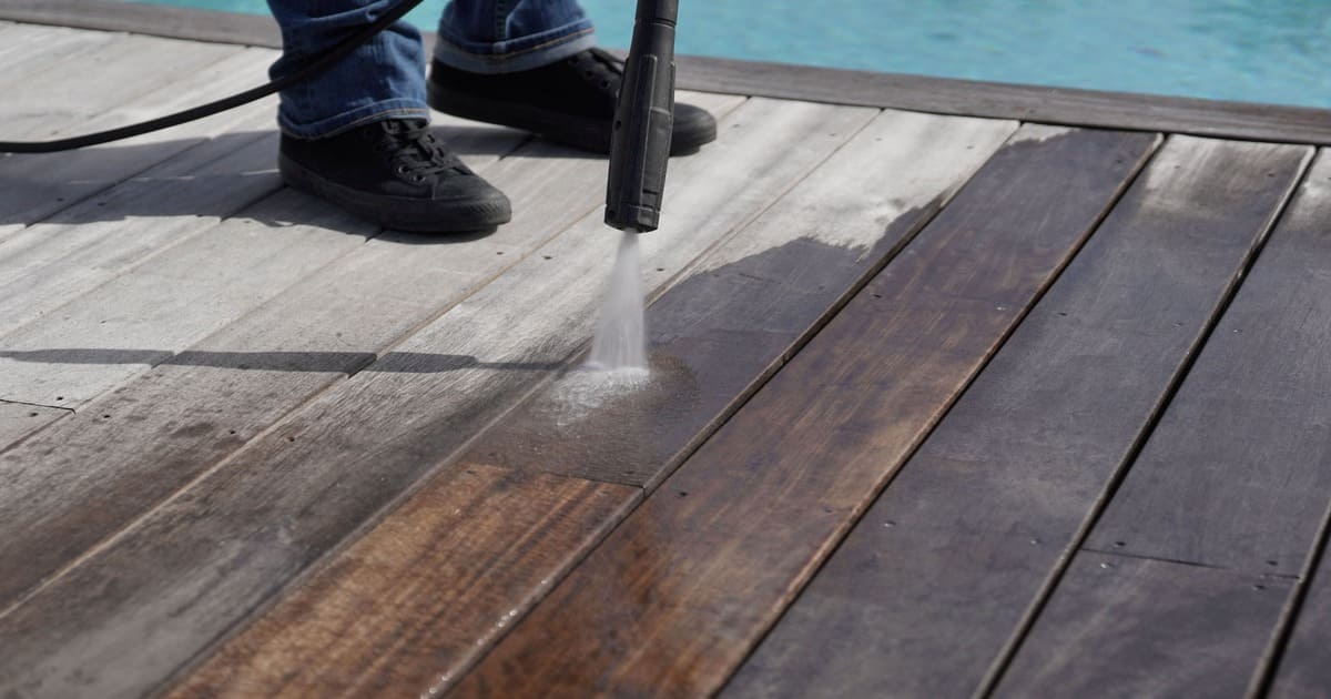 Person using the best deck cleaner and a pressure washer to restore dirty grey wood planks to their original brown finish next to a swimming pool.