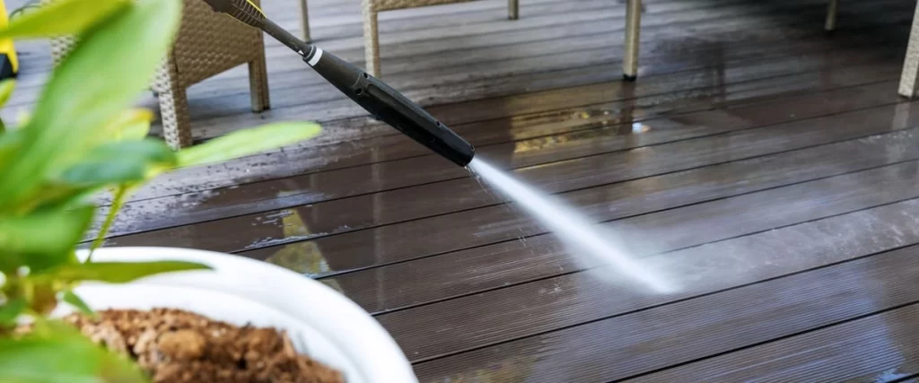 A pressure washer wand spraying a high-powered stream of water onto dark brown grooved deck boards to remove grime and buildup.