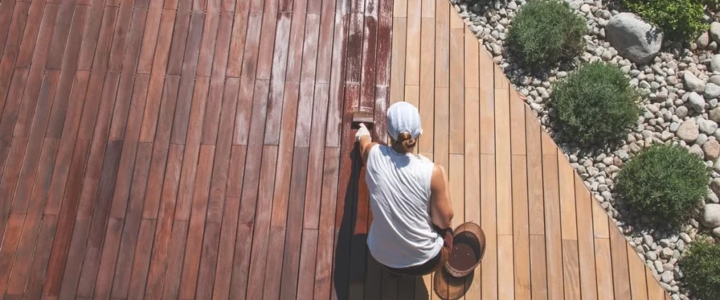 Top-down view of a worker in a white cap hand-painting a large timber deck with a brush and a bucket of reddish-brown stain.