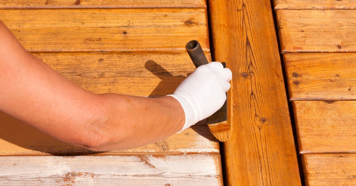 A close-up shot of a hand in a white glove using a brush to apply the best deck stain and sealer to weathered cedar wood boards.