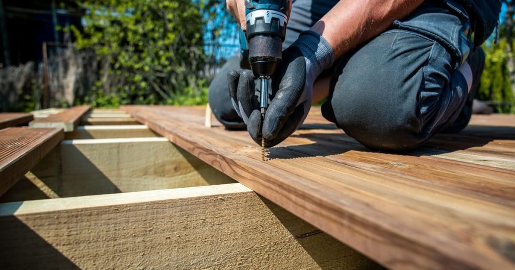 A carpenter using a power drill to secure wooden planks while building a deck around a tree in a sunny backyard.