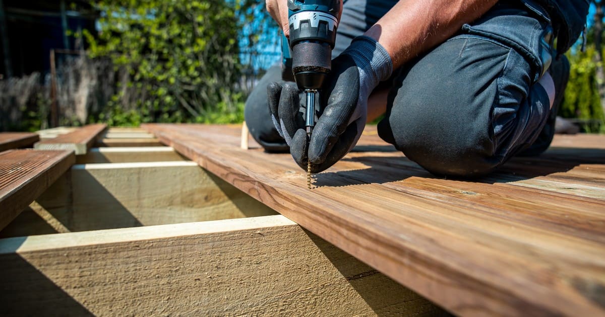 A carpenter using a power drill to secure wooden planks while building a deck around a tree in a sunny backyard.