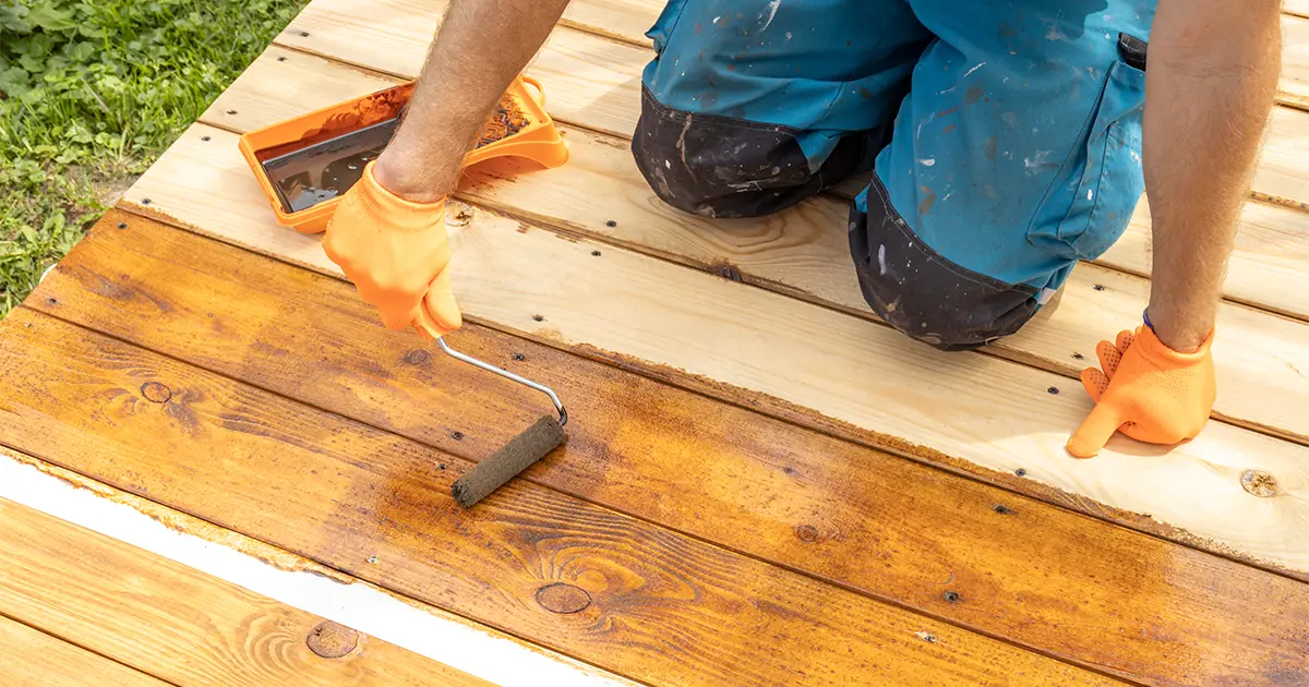 Contractor applying deck sealer with roller on wooden deck boards during deck sealing and maintenance process