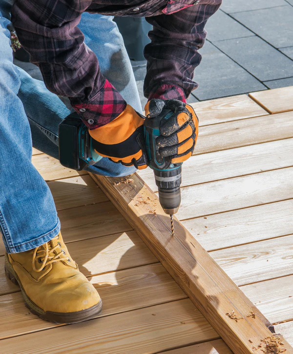 A contractor wearing work gloves and boots uses a cordless drill to secure wooden planks during a deck repair project in Sanford, NC.
